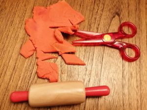 Plastic scissors, toy rolling pin, and a sheet of rolled play-dough with cuts in it.